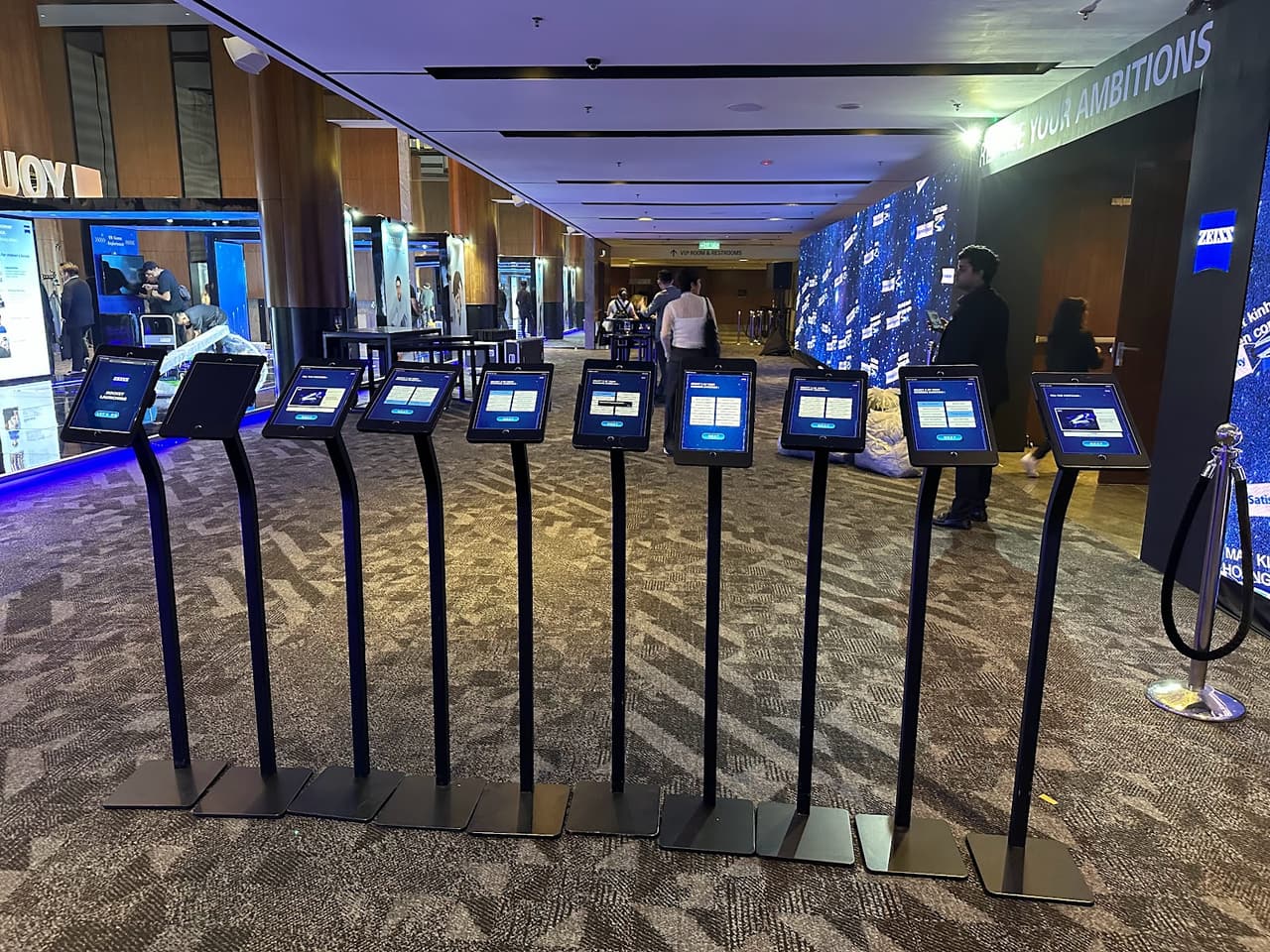 Ten iPad floor stands lined up along an exhibition hall walkway with a lit LED wall behind