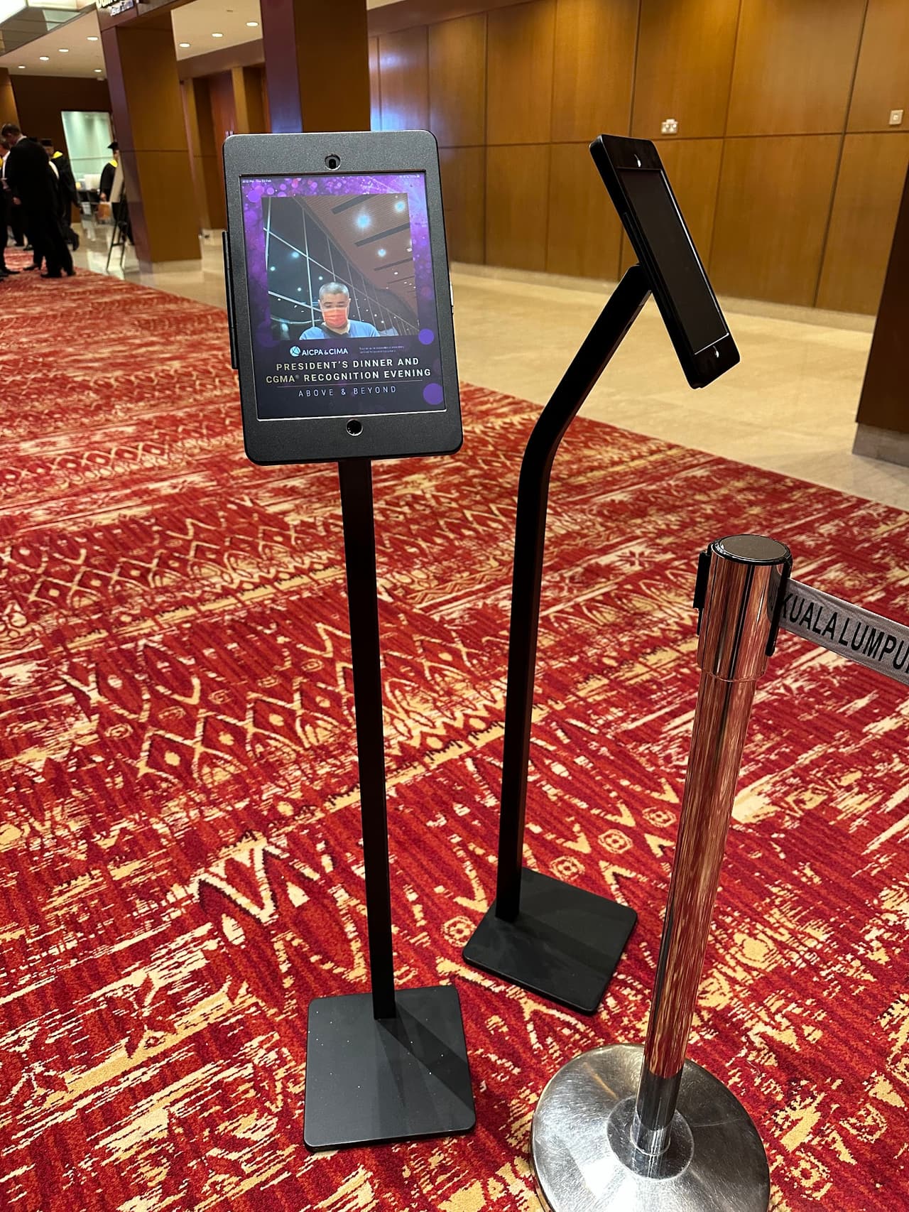 Two iPad floor stands beside a queue barrier on the red carpet of a hotel ballroom foyer
